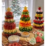 Three food towers shaped like Christmas trees arranged on a large wooden charcuterie board.