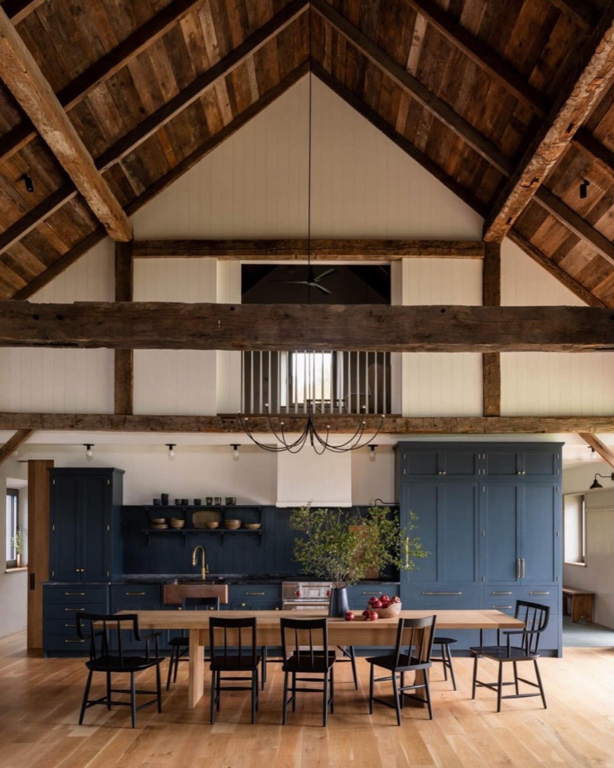 A rustic kitchen with dark blue cabinets, high wooden beamed ceilings, and a long wooden dining table.