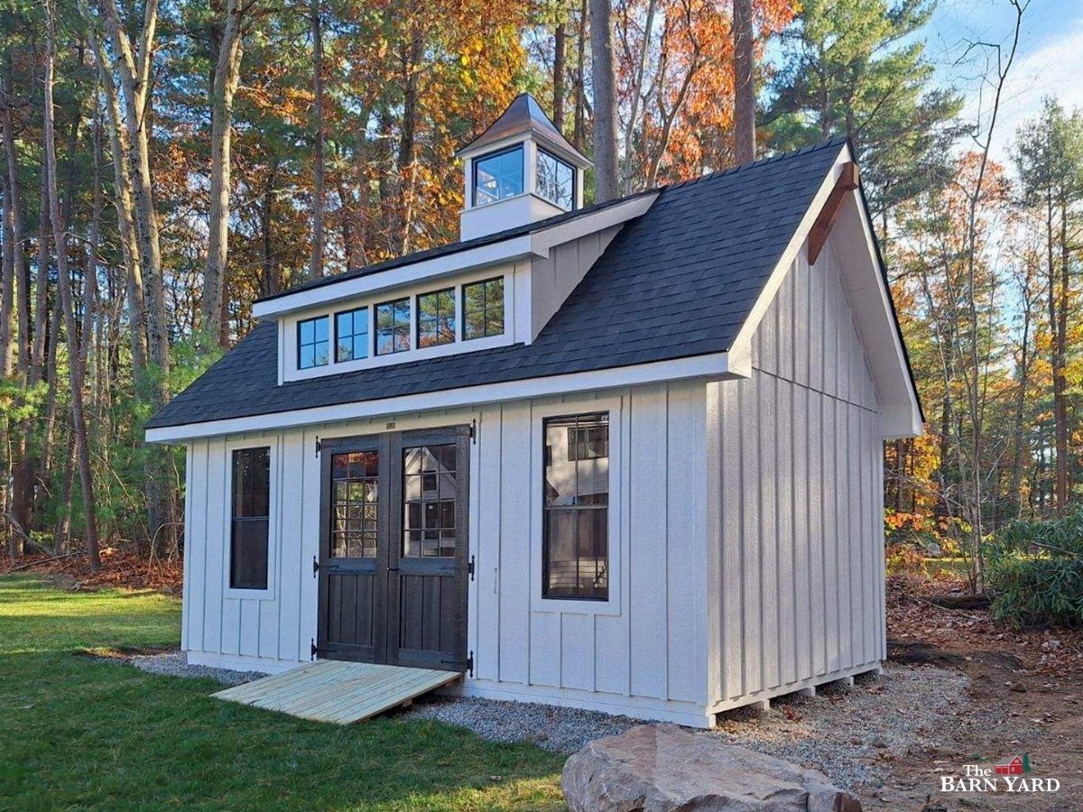 White farmhouse shed with a large shed dormer and cupola in a forest setting.