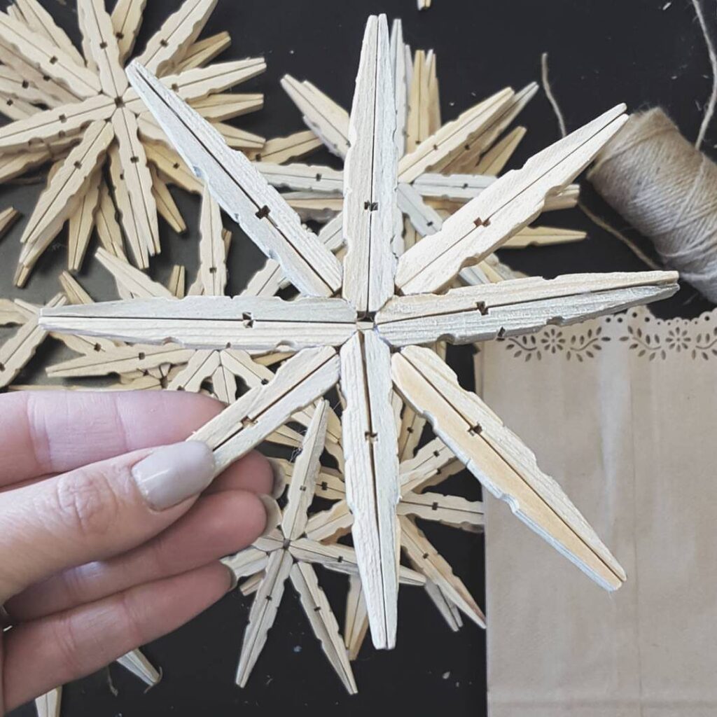 A hand holding a snowflake ornament crafted from multiple light wooden clothespins on a dark background.